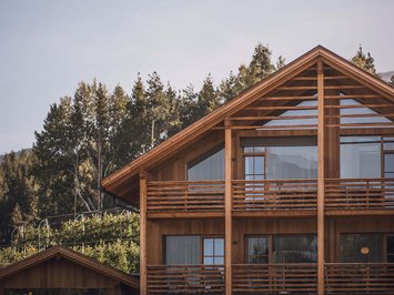 Urlaub in Natz-Schabs? Kessler’s Mountain Lodge! Modernes Holzhaus mit großen Fenstern und Balkon vor Waldkulisse