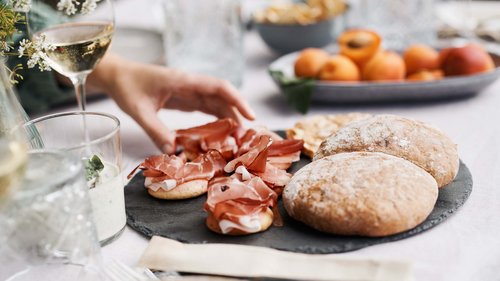 Urlaub in Natz-Schabs? Kessler’s Mountain Lodge! Hand nimmt Prosciutto auf dünnem Brot mit Brötchen und Wein auf gedecktem Tisch