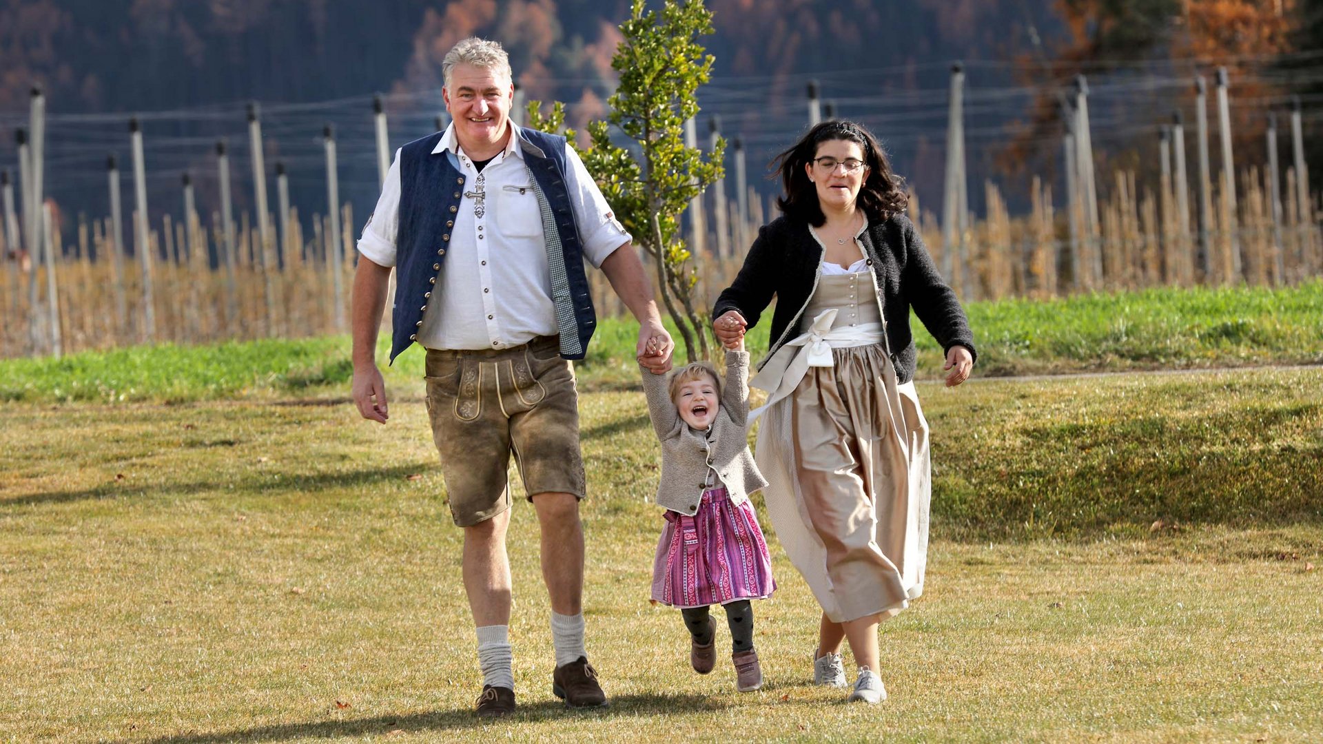 Urlaub in Natz-Schabs? Kessler’s Mountain Lodge! Glückliche Familie in traditioneller Kleidung läuft im Grünen und hält Kind an den Händen
