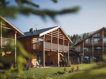 Urlaub in Natz-Schabs? Kessler’s Mountain Lodge! Holzchalets mit Balkonen und Liegestühlen auf grünem Rasen bei sonnigem Wetter
