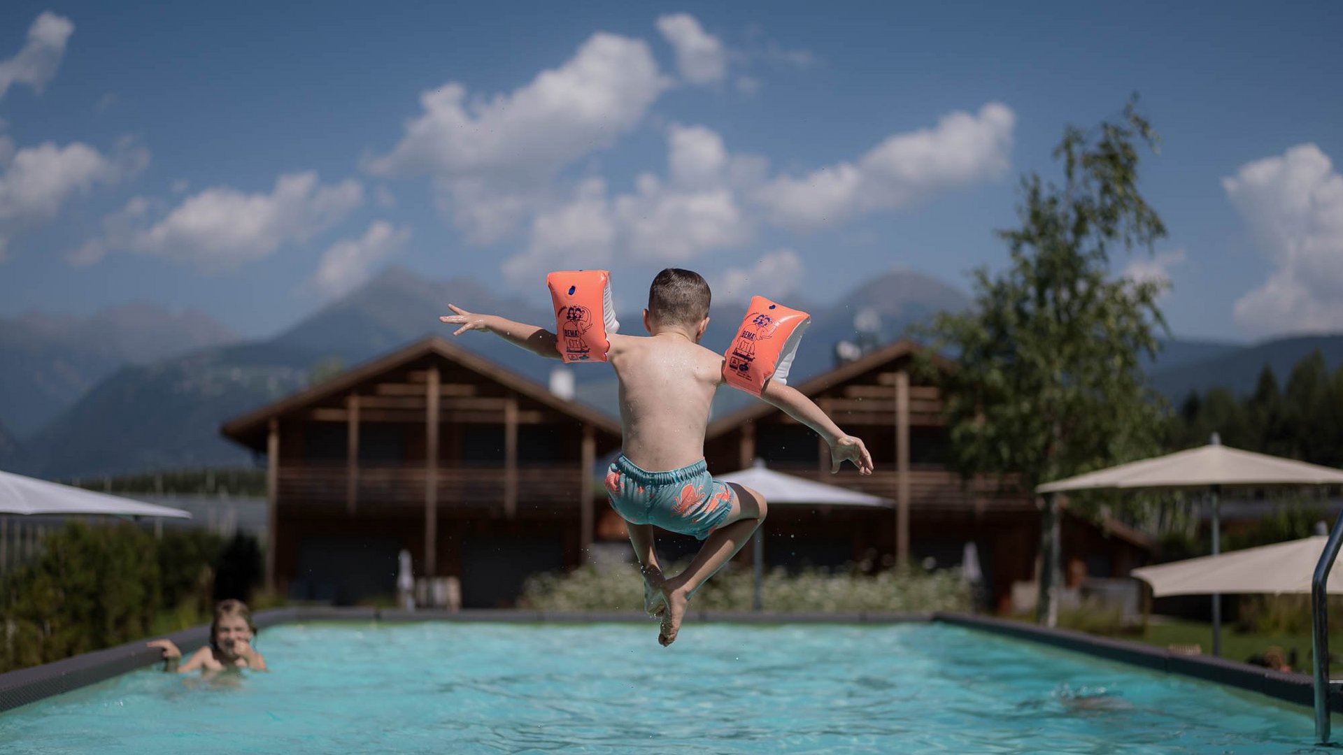 Urlaub in Natz-Schabs? Kessler’s Mountain Lodge! Kind mit Schwimmflügeln springt in einen Außenpool vor Berghütten